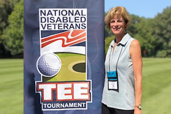 Deborah Voydetich stands next to a banner that says National Disabled Veterans Tee Tournament.