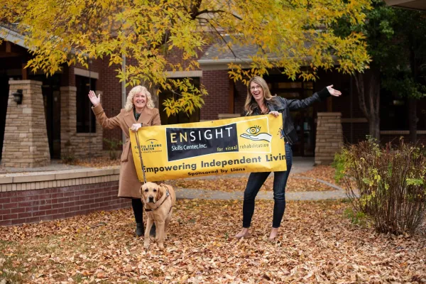 Two women hold a banner with the words Ensight Skills Center, Inc. A golden retriever stands in front of the woman on the left.