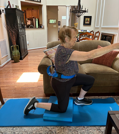 A elderly woman does stretching exercises in a half kneeling position.