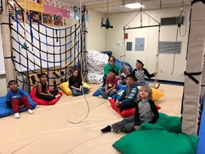 A semicircle of eight children sit on cushions in a therapy gym. An adult woman sits in the background.